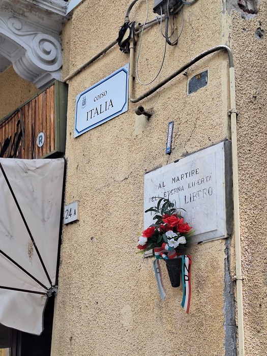 Lapide commemorativa su una parete di un edificio, con fiori e decorazione tricolore.