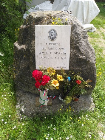 Cippo commemorativo dedicato al partigiano Amleto Grazia, decorato con fiori e un omaggio alla Resistenza.