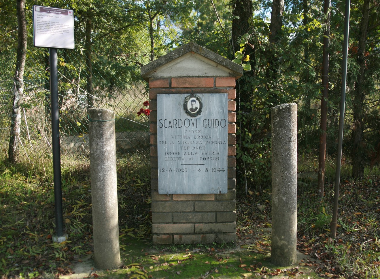 Cippo commemorativo dedicato a Scardovi Guido, immerso in un contesto naturale con alberi e recinzione.