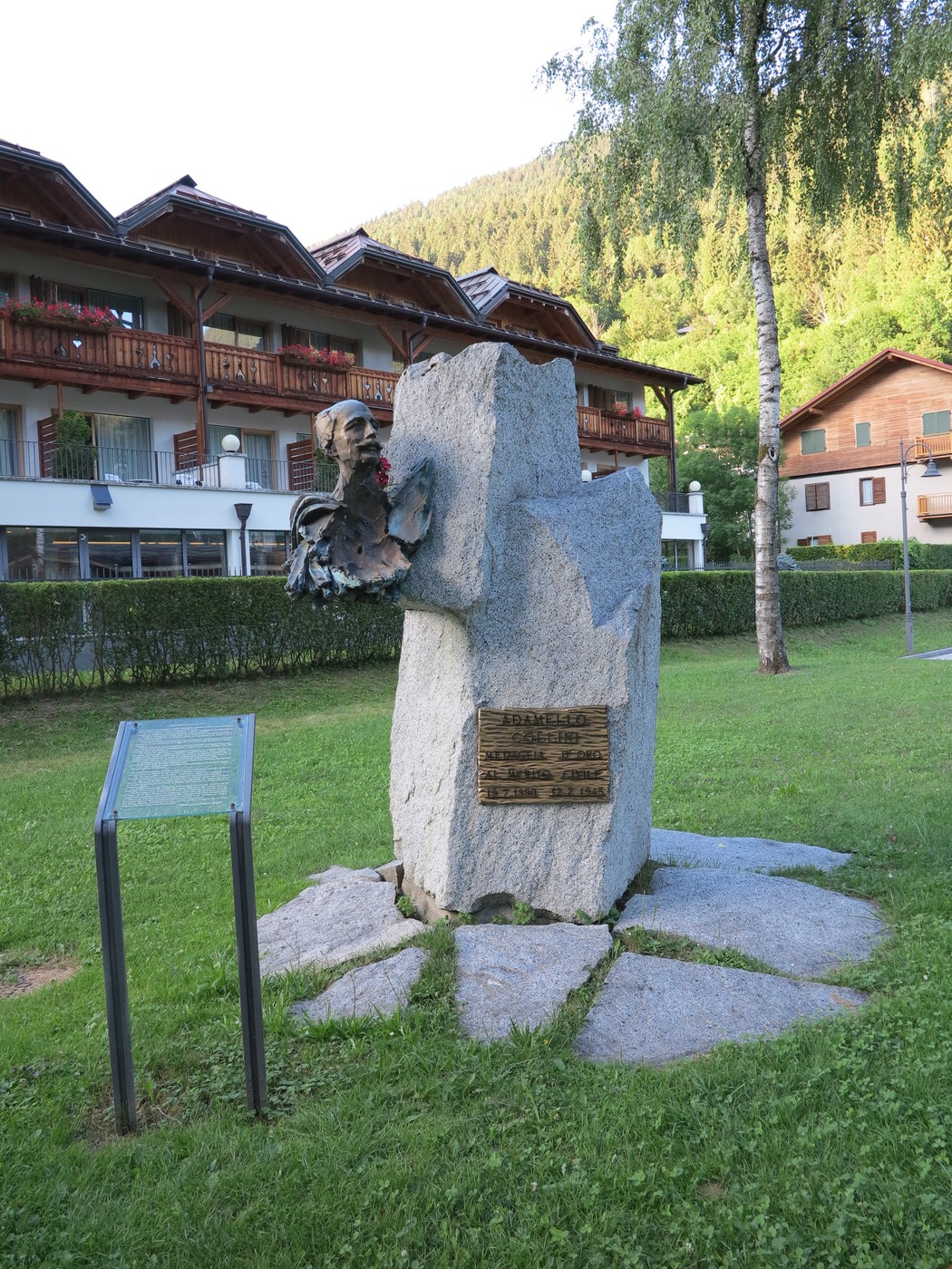 Monumento a Collini, scultura in pietra con busto e pannello descrittivo, circondato da verde e edifici.