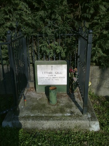 Cippo commemorativo dedicato a Ettore Gilli, con fiori e una recinzione metallica.