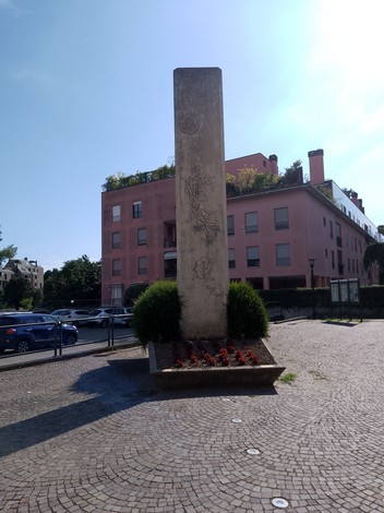 Monumento alla Resistenza, obelisco decorato, circondato da aiuole fiorite e edifici residenziali.