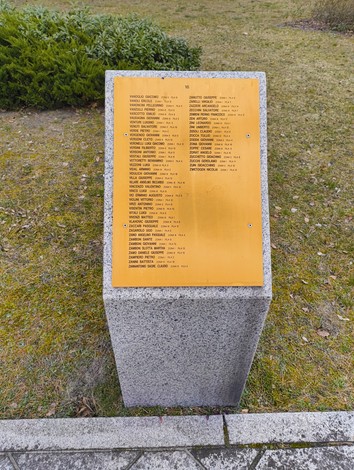 Pannello commemorativo con nomi di caduti nel Cimitero militare italiano di Zehlendorf, circondato da verde.