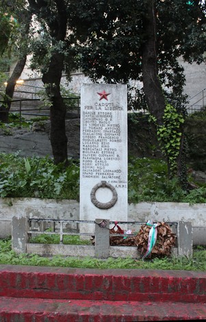 Monumento ai caduti per la Libertà di Staglieno, circondato da vegetazione e decorato con una corona in memoria.