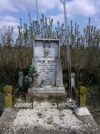 Cippo commemorativo dedicato a Saccani Antonio, immerso nella natura con fiori e simboli di memoria.