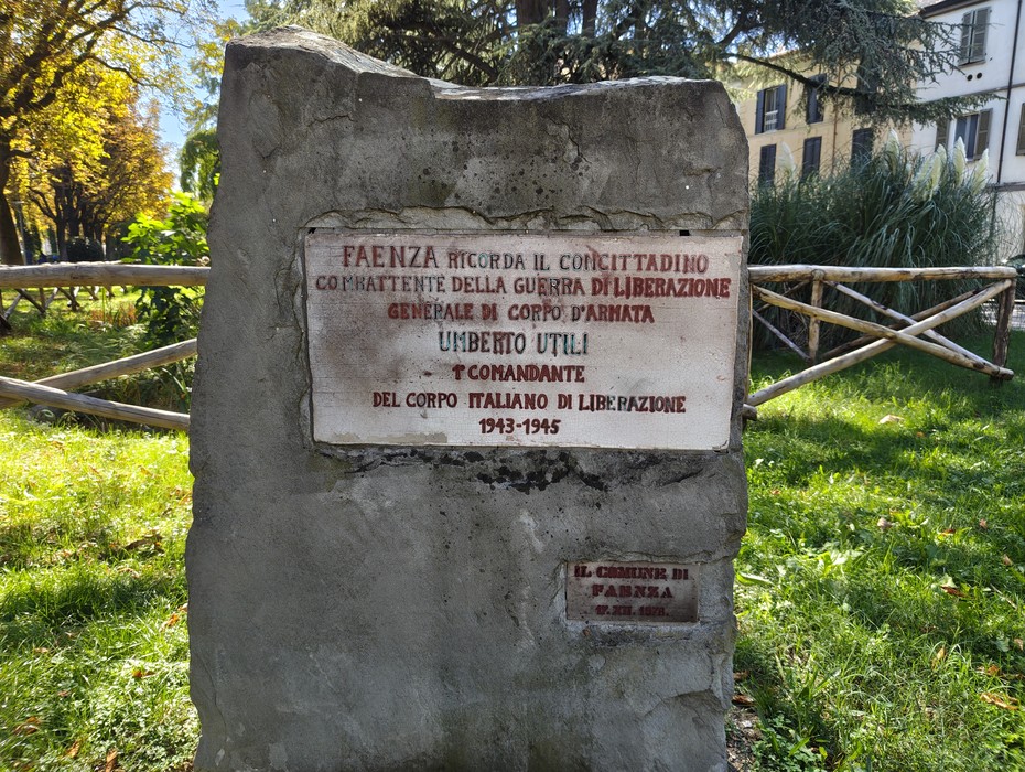Cippo in pietra con iscrizione e targhetta, collocato in un giardino tra alberi e staccionate di legno.
