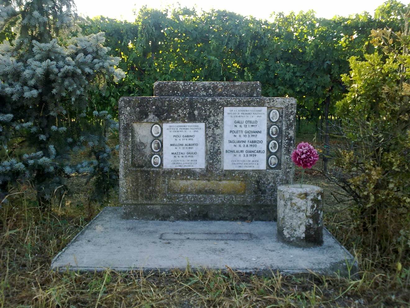 Lapide commemorativa a Bonilauri con nomi e dati di persone ricordate, circondata da vegetazione.