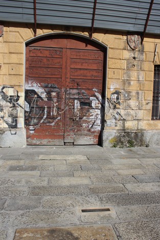Soglia d'entrata con porta in legno scuro e pareti in pietra, segni di usura e graffiti visibili.