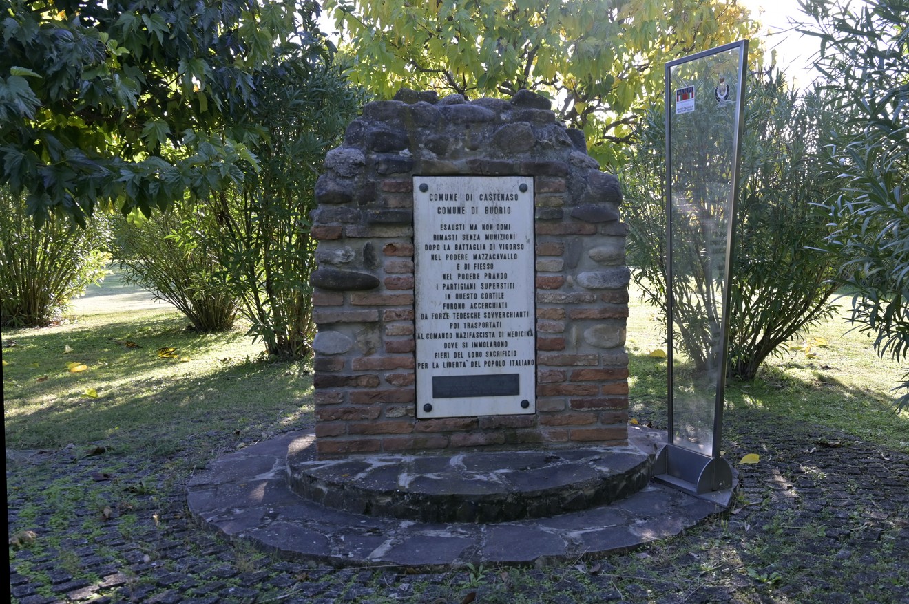 Monumento commemorativo alla battaglia di Casa Mengoli, circondato da vegetazione, con una lapide esplicativa.