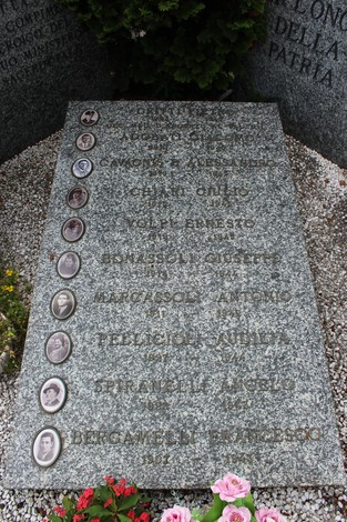 Targa commemorativa con nomi e fotografie dei caduti per la patria, circondata da fiori e piante.