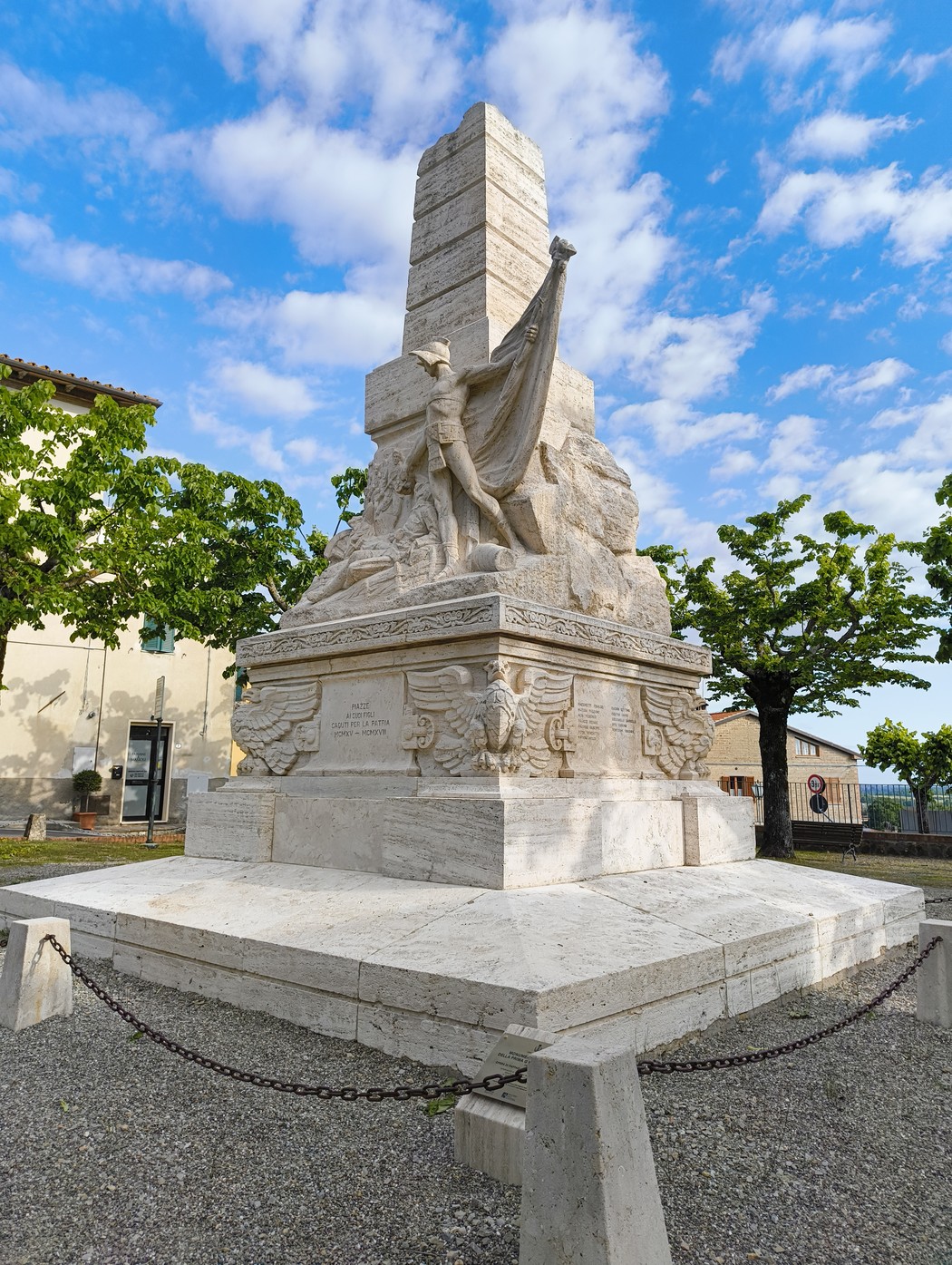 Monumento ai caduti di Piazze, con statua centrale e base monumentale, circondato da alberi e cielo azzurro.