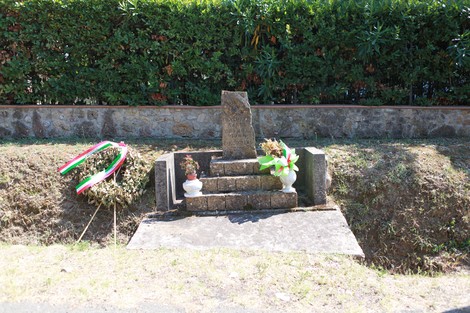 Cippo commemorativo dedicato a Fantozzi, con fiori e decorazioni tricolori nel contesto naturale.