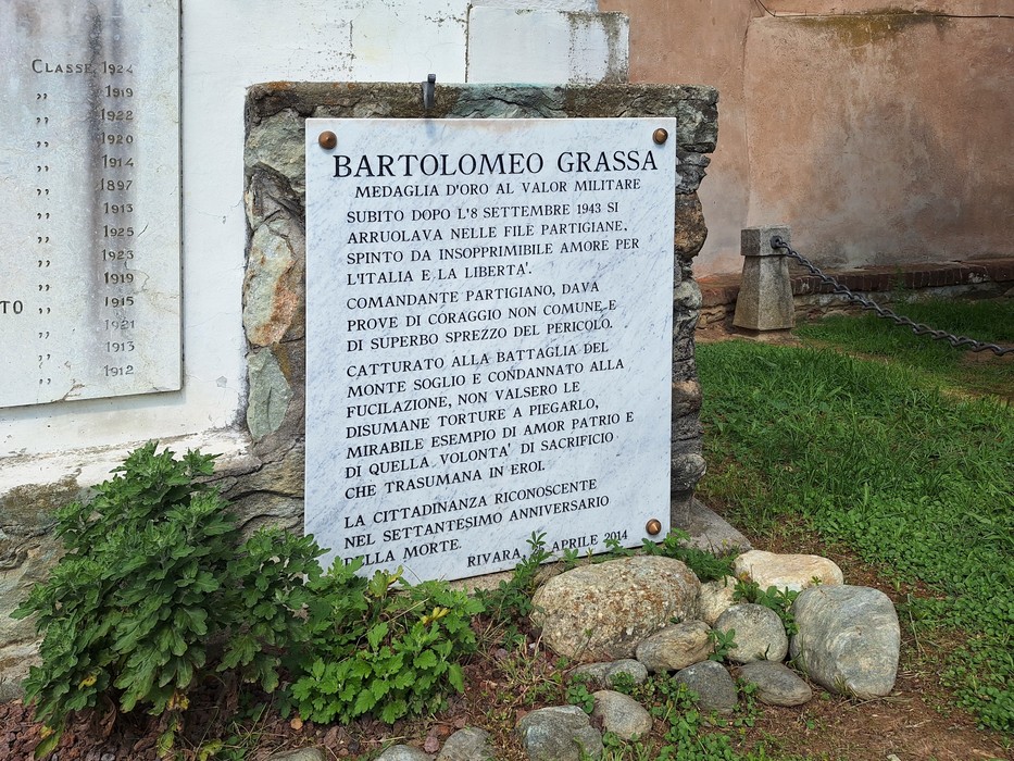 Lapide in pietra con iscrizione, fissata a una parete esterna e circondata da erba e pietre.