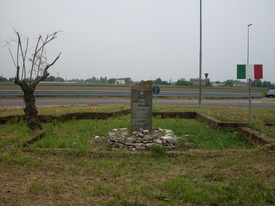 Cippo commemorativo immerso nel verde, con strada e paesaggio al fondo. Atmosfera di riflessione e memoria.