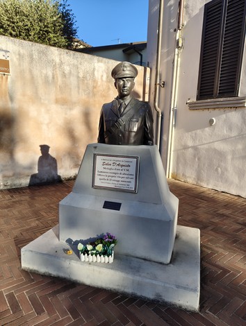 Monumento a D'Acquisto, con statua e base commemorativa, circondato da fiori in una piazza soleggiata.