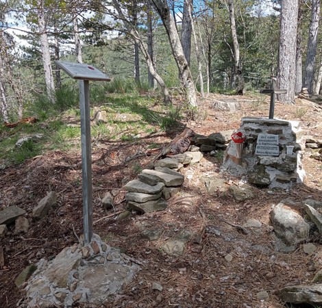 Cippo commemorativo in un'area boschiva, circondato da pietre e segnato da due croci. Atmosfera tranquilla e naturale.
