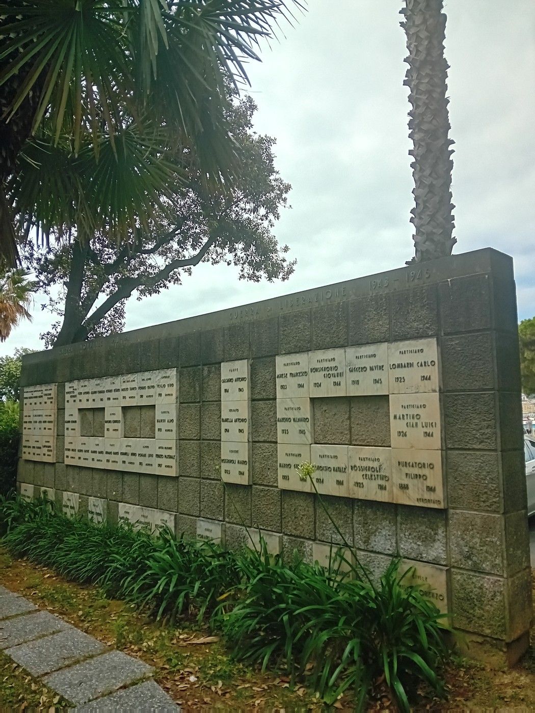 Monumento ai caduti, stone wall with engraved names, surrounded by greenery and palm trees.