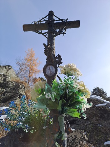 Croce in ferro con corona di spine, fiori artificiali e orologio, tra rocce e alberi.