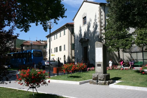 Cippo commemorativo ai caduti, circondato da fiori e edifici, in un tranquillo spazio verde.