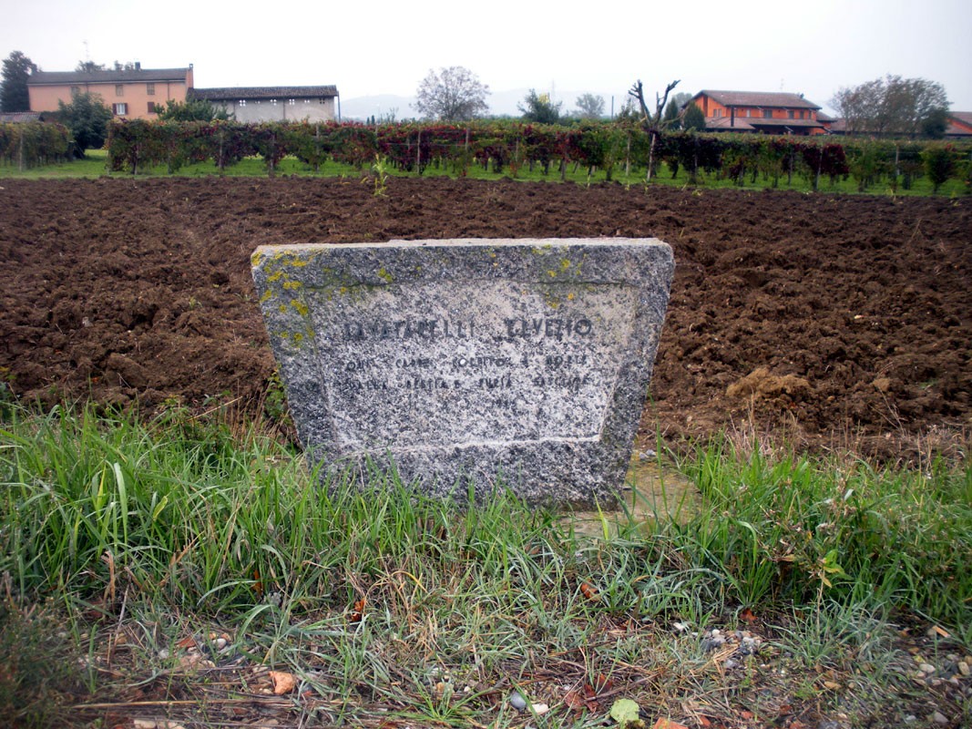 Lapide commemorativa a Zavattarelli, situata in un campo circondato da vegetazione e edifici rurali.