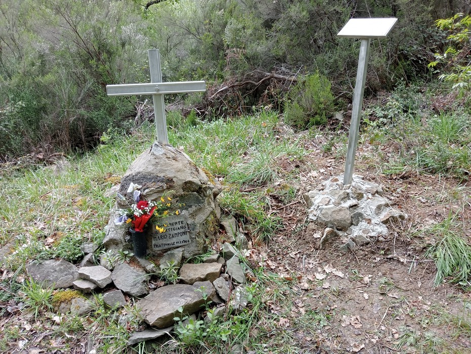 Cippo commemorativo a Zamponi, con fiori e croce, immerso nella natura. Sentiero verde circostante.