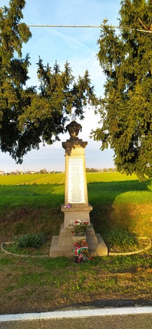 Monumento ai caduti dell'Amola immerso in un paesaggio rurale, con alberi e un campo sullo sfondo.