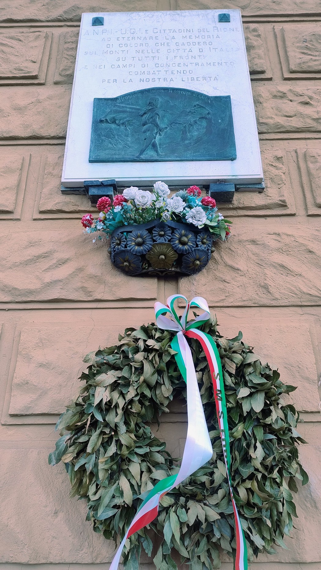 Lapide commemorativa dedicata ai caduti, adornata con una corona di alloro e fiori, su un muro di pietra.