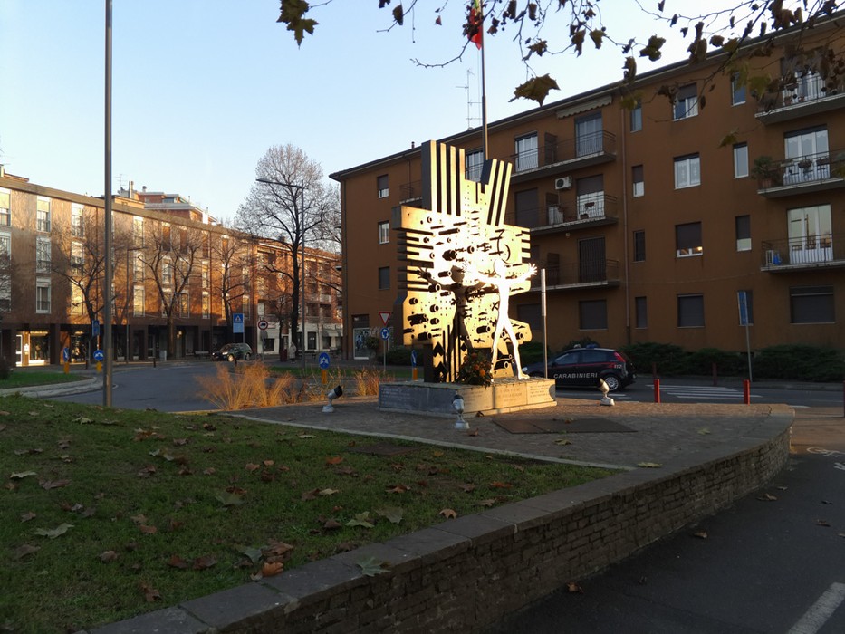 Monumento metallico astratto su piazza urbana, circondato da edifici e alberi spogli.
