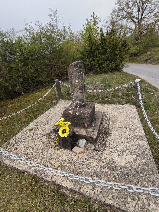 Cippo commemorativo a Ulivelli circondato da catene e fiori, immerso nella natura.
