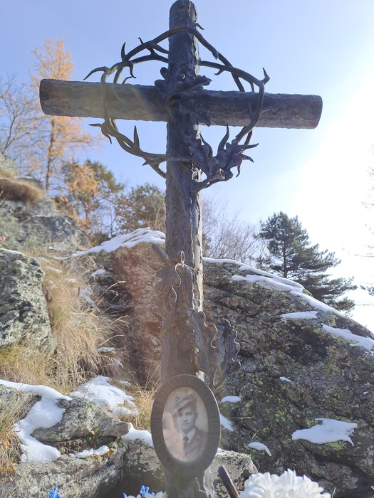Croce in legno con corona di rami metallici intrecciati, tra rocce e vegetazione, con foto ovale di un uomo in divisa.