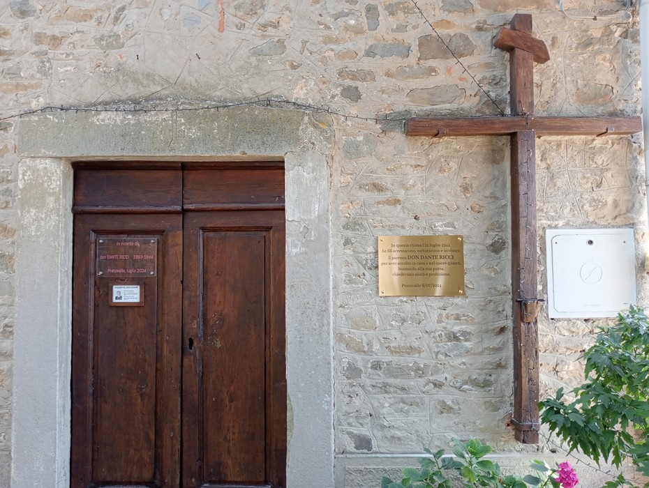 Porta in legno e croce di legno su parete di pietra, con targa informativa accanto. Atmosfera storica e rustica.