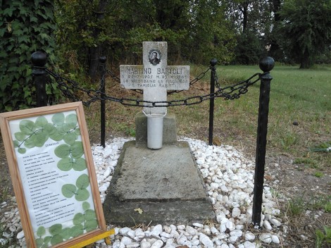 Monumento dedicato a Bartoli, con croce e decorazioni floreali, immerso in un ambiente naturale.