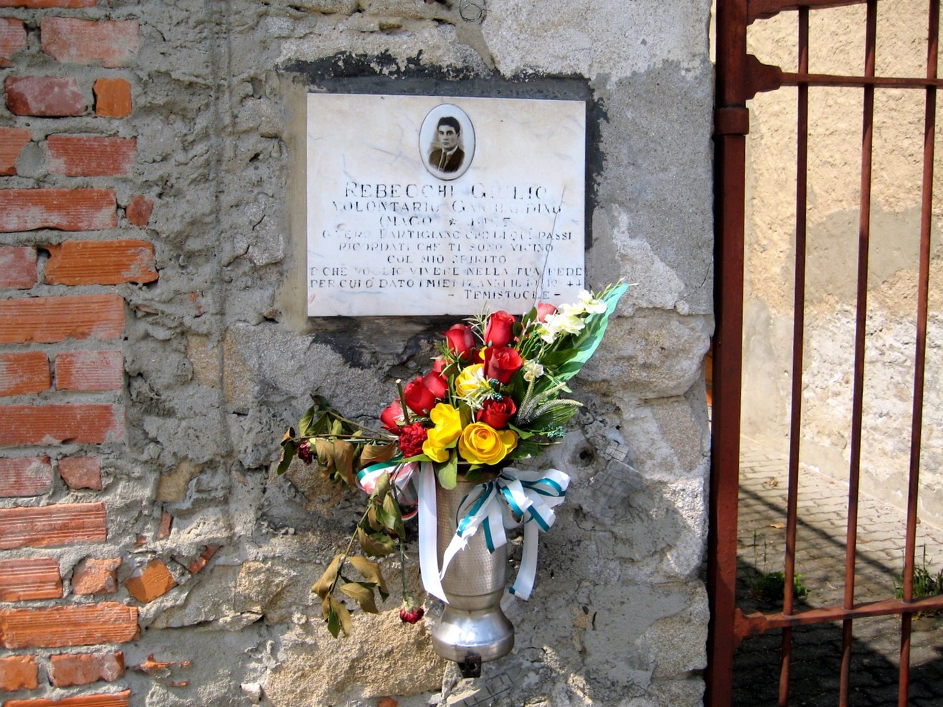 Lapide commemorativa di Rebecchi, adornata con fiori, su un muro di pietra.