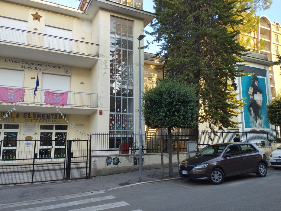 Murale agli Eroi ottobrini su un edificio scolastico, con decorazioni colorate alle finestre e una bandiera esposta.