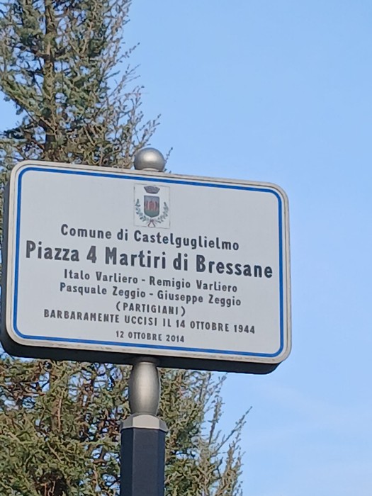 Cartello stradale commemorativo con iscrizione, montato su un palo davanti ad alberi e cielo.