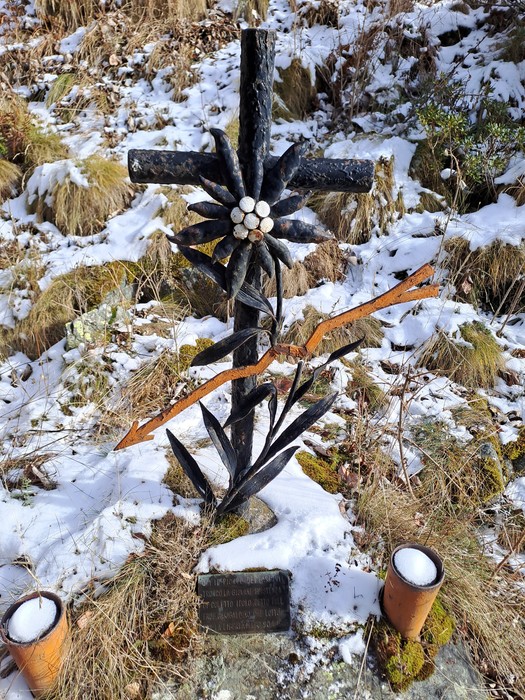 Croce in ferro battuto con stella alpina decorativa e targa, su basamento tra erba e neve nel bosco.