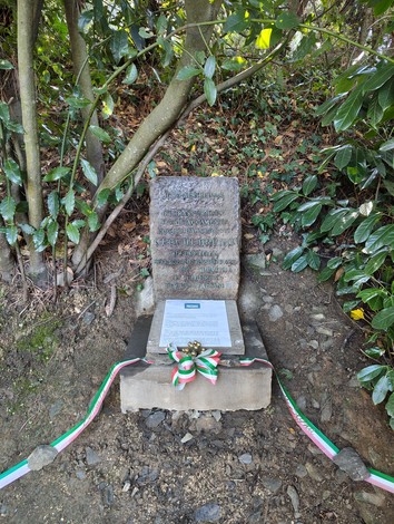 Cippo commemorativo circondato da vegetazione, con una corona tricolore davanti e iscrizioni sulla pietra.