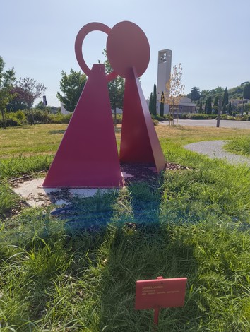Scultura rossa che rappresenta due figure stilizzate, circondata da un verde prato e con luce solare.