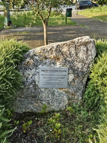 Monumento a Fusconi, una stele in pietra con una placca commemorativa circondata da vegetazione.