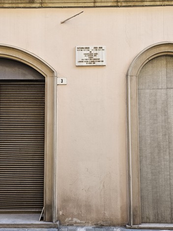 Lapide commemorativa su muro in un edificio, sovrastata da due archi, uno con serranda e l'altro chiuso.