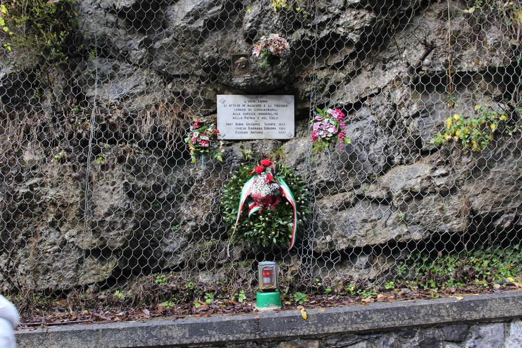 Lapide commemorativa adornata da fiori su una parete rocciosa in un contesto naturale.