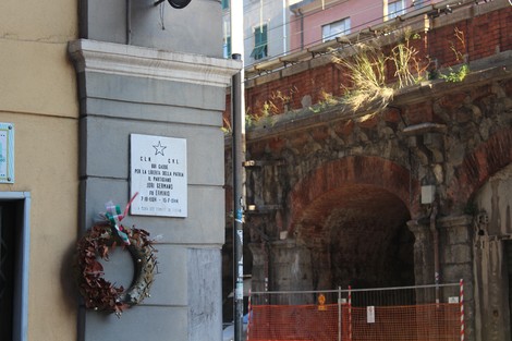 Lapide commemorativa con decorazioni natalizie, situata in un contesto urbano, vicino a archi in pietra.
