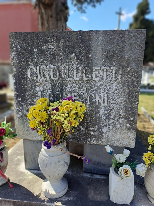 Monumento a Lucetti adornato con fiori, situato in un cimitero circondato da alberi e un cielo sereno.