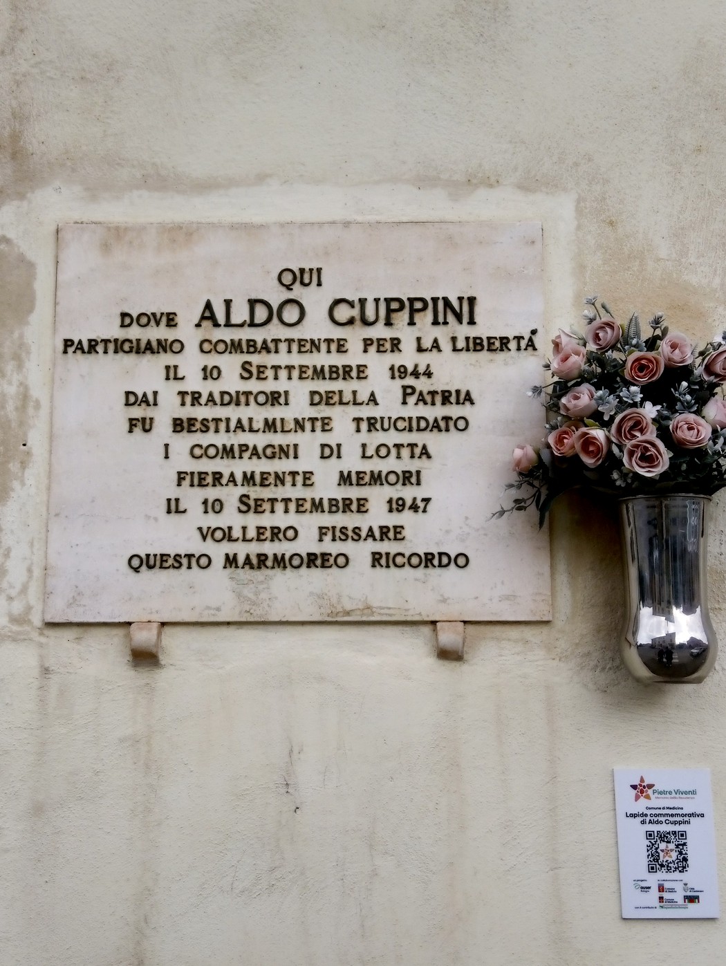 Lapide commemorativa dedicata ad Aldo Cuppini, partigiano, situata su una parete, circondata da fiori artificiali.