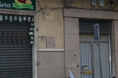 Lapide commemorativa su un edificio, visibile tra un'insegna e una finestra, in un contesto urbano.