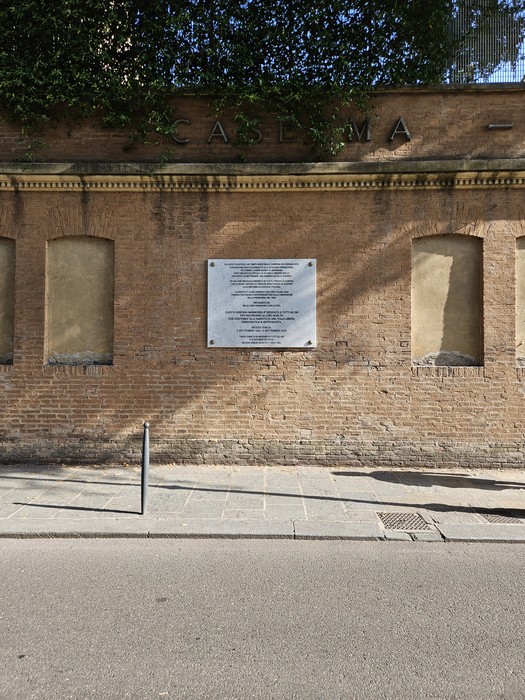 Lapide in marmo con iscrizione, affissa su una parete in mattoni con la scritta "Caserma".