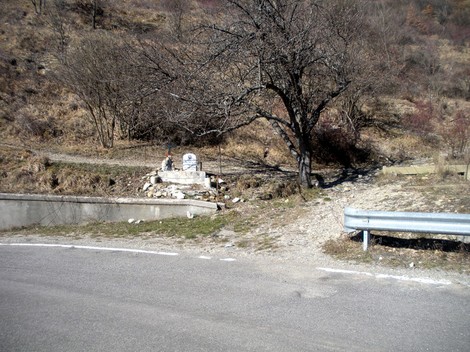 Cippo commemorativo su strada, circondato da natura e piante, in una zona montana.