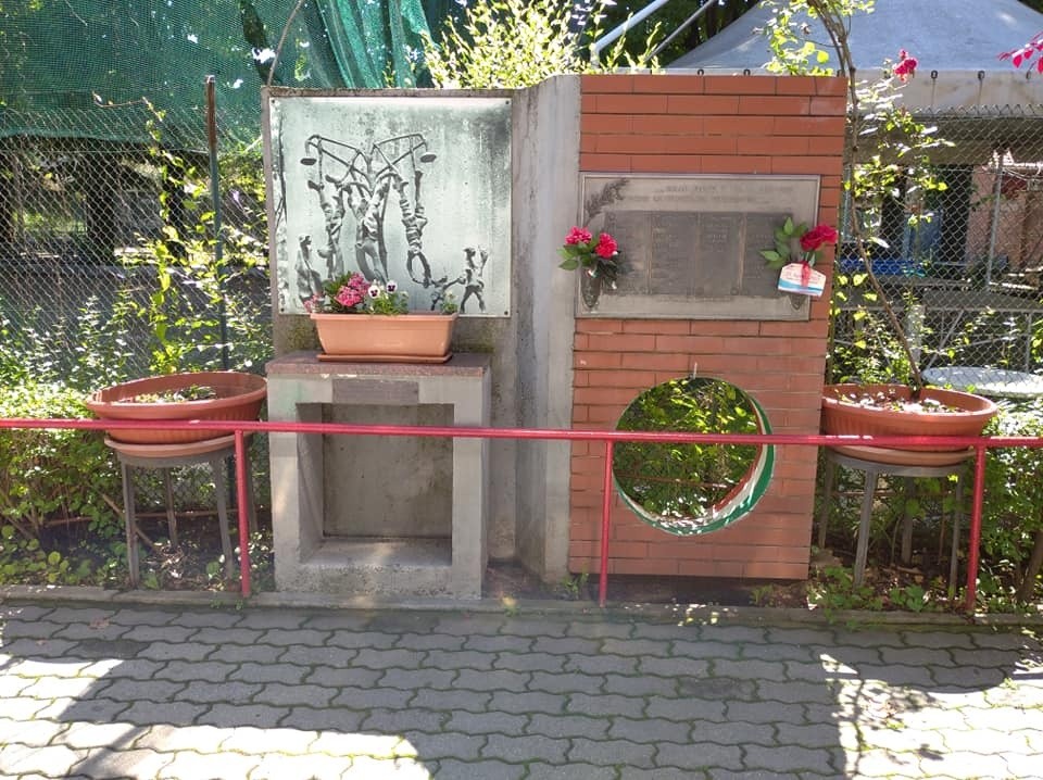 Monumento ai caduti con elementi commemorativi e decorazioni floreali in un contesto verde.