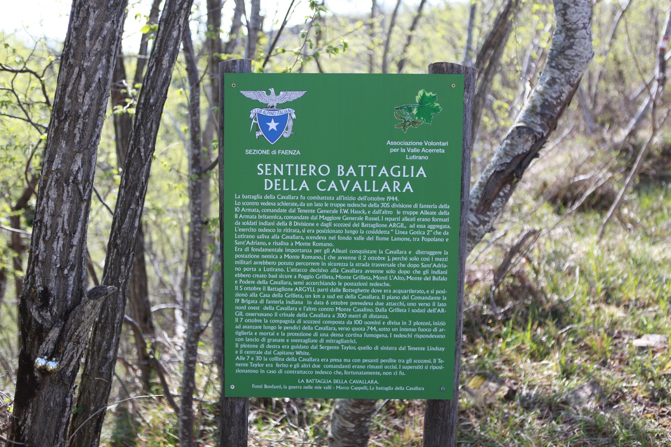 Pannello informativo sul "Sentiero Battaglia della Cavallara" immerso nella natura, circondato da alberi e vegetazione.