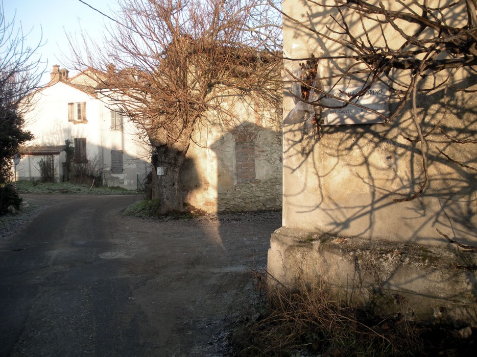 Immagine di un angolo di un villaggio con edifici e ombre di alberi, rappresenta "Lapide a Bigoni".